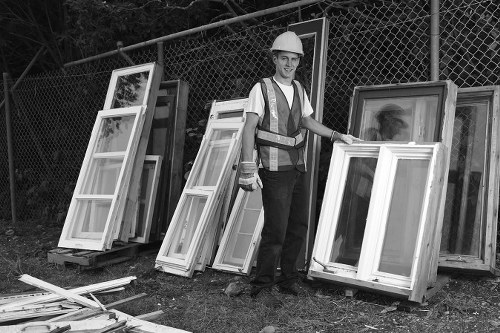 A construction worker wearing a white safety helmet and a high-visibility vest stands outdoors on a grassy area near a chain-link fence, next to a stack of white window frames with glass panes. The frames, which vary in size, are leaning against the fence and placed on the ground, some arranged upright and others lying flat. The worker is smiling and holding one of the window frames, indicating involvement in a building or refurbishment project, possibly in Ilford or nearby. The background includes trees and a partly cloudy sky, suggesting daytime conditions. This scene relates to rubbish removal and clearance services for building waste and refurbishment debris in the local area, aligning with professional waste management practices.