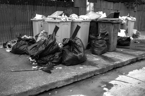 A row of four large wheelie bins positioned on a paved driveway area next to a wooden fence, with the bins filled with various types of rubbish. The first two bins from the left are light grey, the third bin is dark grey, and the fourth bin appears to be black or dark brown. Several garbage bags, predominantly black, are placed on the ground in front of and around the bins, some tied and others open with visible waste spilling out. The rubbish includes crumpled paper, plastic wrappers, and miscellaneous waste materials. The surface beneath the bins and bags is concrete, with some debris scattered nearby, indicating an area used for waste disposal or rubbish collection. The background shows a wooden fence, and the overall environment suggests a typical outdoor refuse area in a residential or commercial setting in or near Ilford. This scene is consistent with rubbish removal services operated by Office Clearance Ilford in the local area.