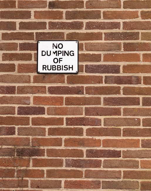 A rectangular white metal sign with black lettering is mounted on a reddish-brown brick wall. The sign displays the message 'NO DUMPING OF RUBBISH,' with the words 'NO DUMPING' positioned above 'OF RU