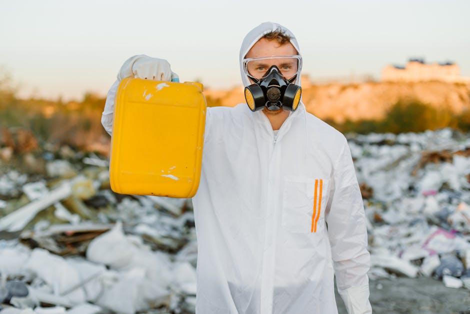 A person wearing a white protective suit, including a hood, safety goggles, and a black respiratory mask, is standing amidst a large pile of mixed waste and rubbish, which includes plastic bottles, ca