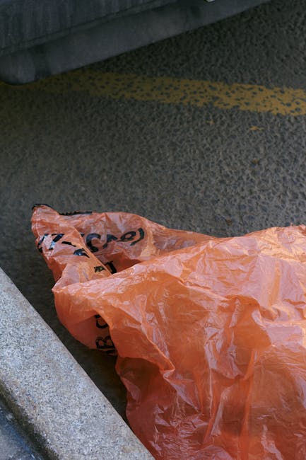 A crumpled orange plastic rubbish bag lying on the asphalt pavement just beside a curb edge in an outdoor urban environment. The bag's material appears thin and slightly reflective, with visible creas
