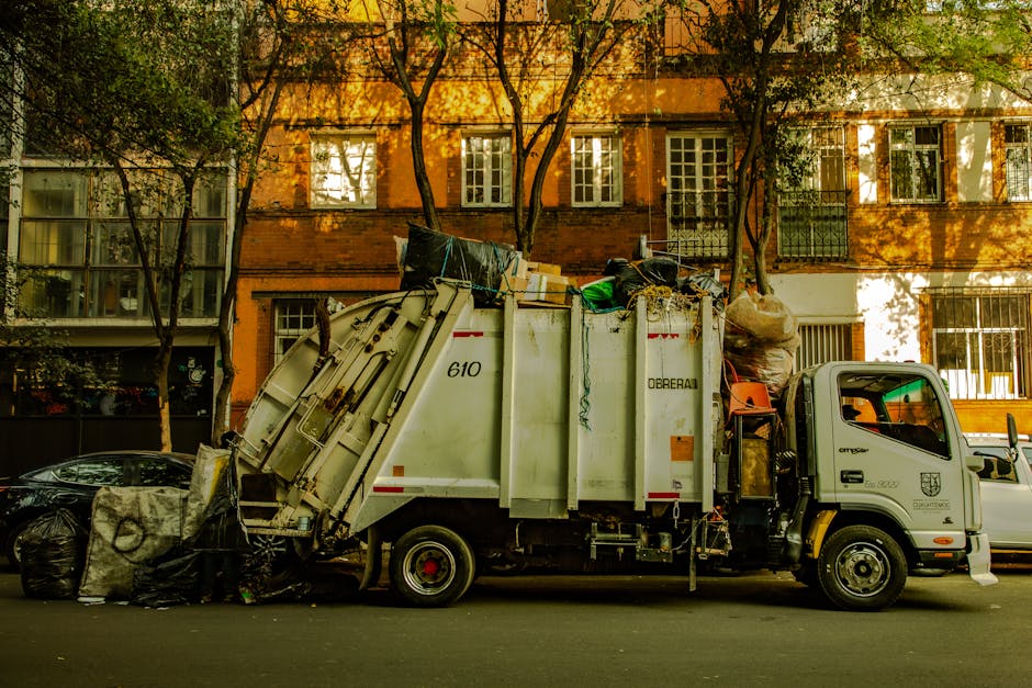A small, dark red flatbed truck parked on an urban street in front of a modern office building with large glass windows and concrete facade. The truck's open cargo bed is filled with various waste mat